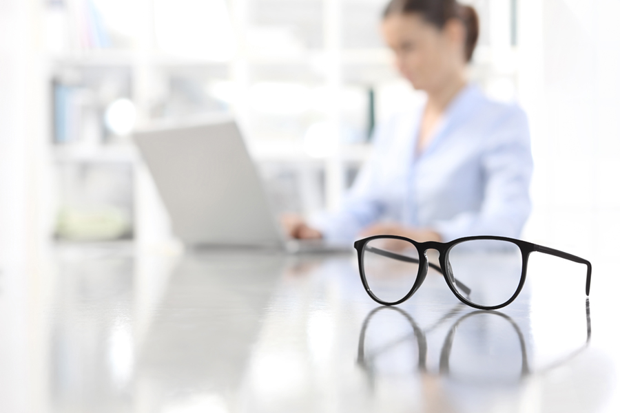 eyeglasses on desk in foreground with blurry woman in background